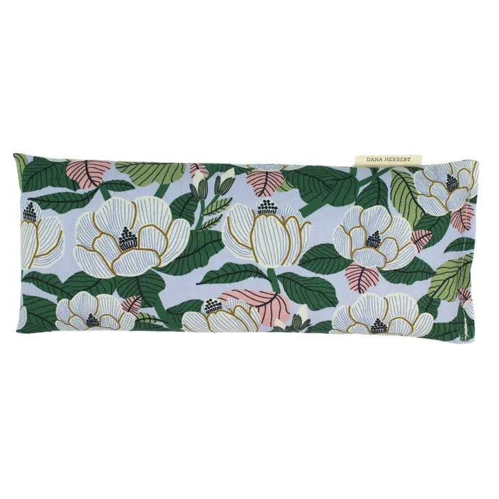 An eye pillow with a botanical pattern of large white magnolia flowers with pink veins on a light blue background. A tag says "Dana Herbert".