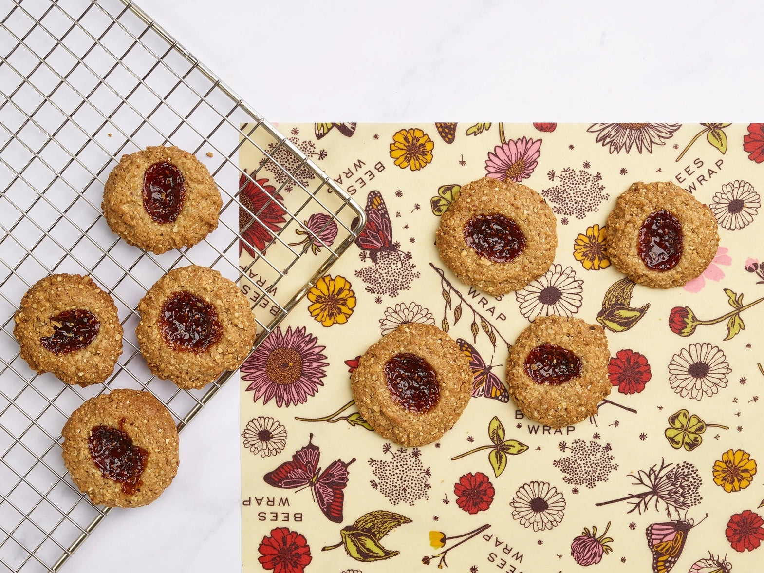 Cookies with jam centers cooling on a rack and placed on Bees Wrap with red and yellow floral and butterfly design.