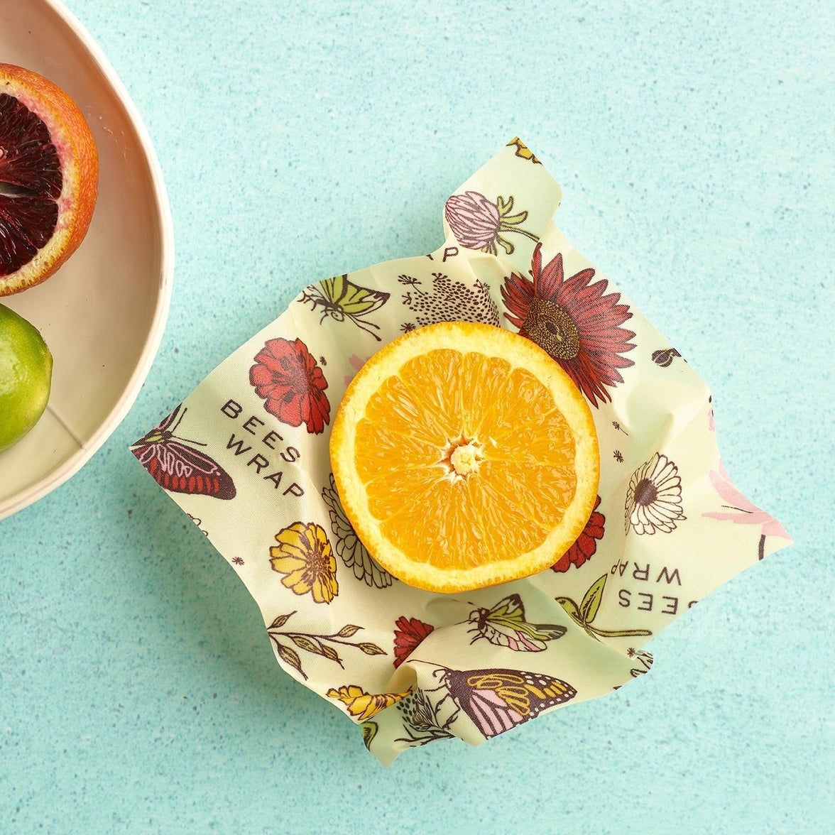 Half of a fresh orange sits on top of a beeswax food wrap decorated with colorful butterflies and flowers, placed on a light blue surface next to a bowl containing a blood orange and a lime.