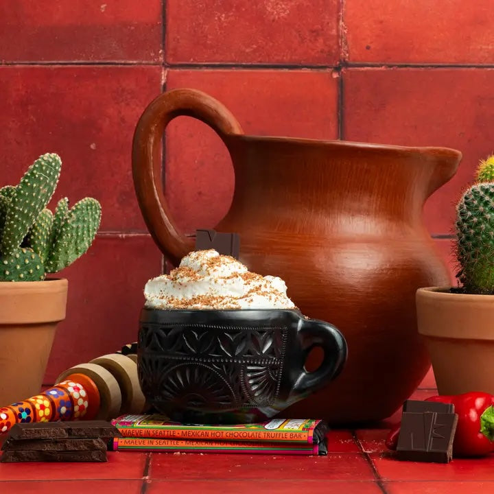 A rustic cup filled with rich Mexican hot chocolate topped with foam, placed on a wooden surface with chocolate pieces nearby, evoking warm, spicy flavors.