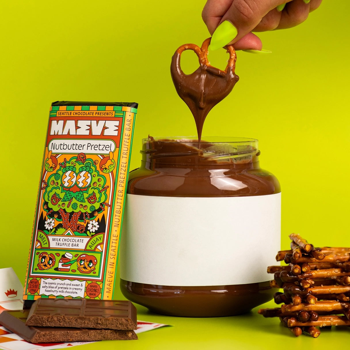 Pieces of the Maeve “Nutbutter Pretzel” chocolate bar next to a nut butter jar with a pretzel dipping into it.