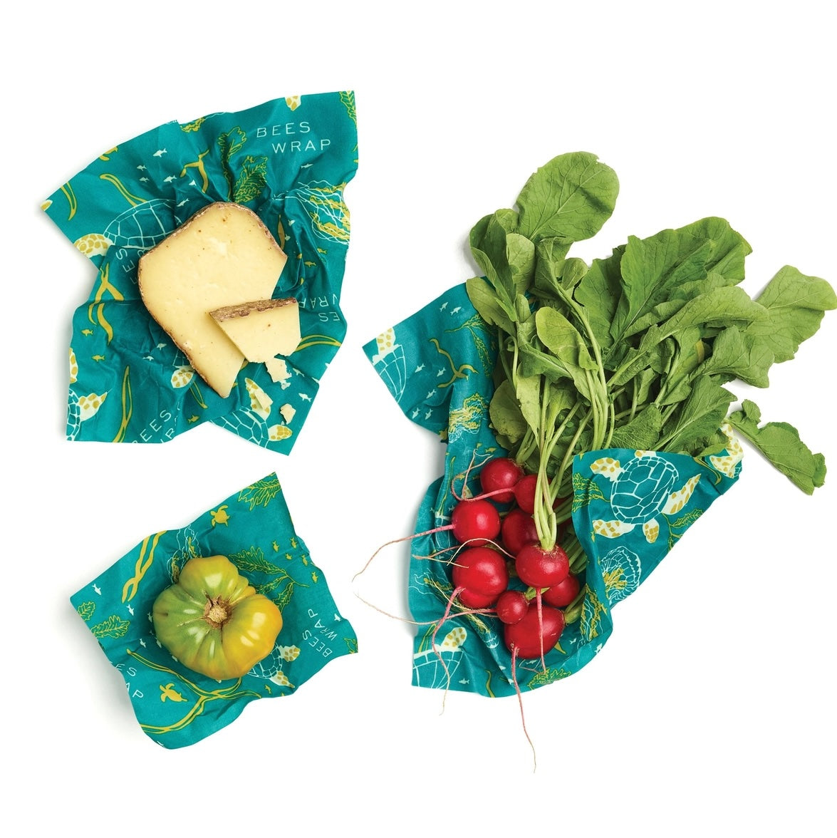Green and yellow heirloom tomato, cheese wedge, and radishes wrapped in teal beeswax wrap with sea turtle designs.