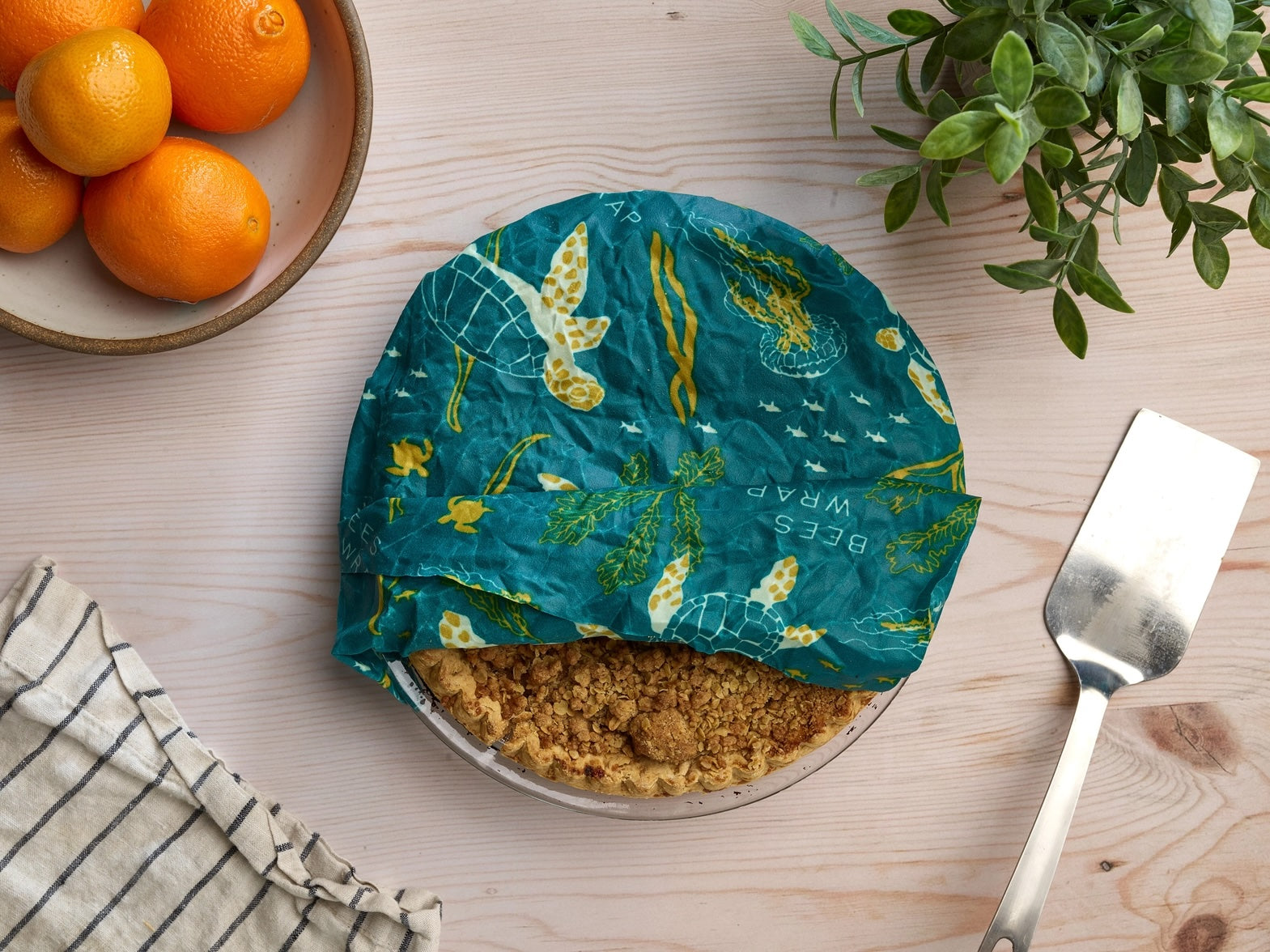 Top view of a teal-colored beeswax food wrap with marine life and seaweed designs covering a pie, next to a metal pie server and cloth napkin on a wooden table.