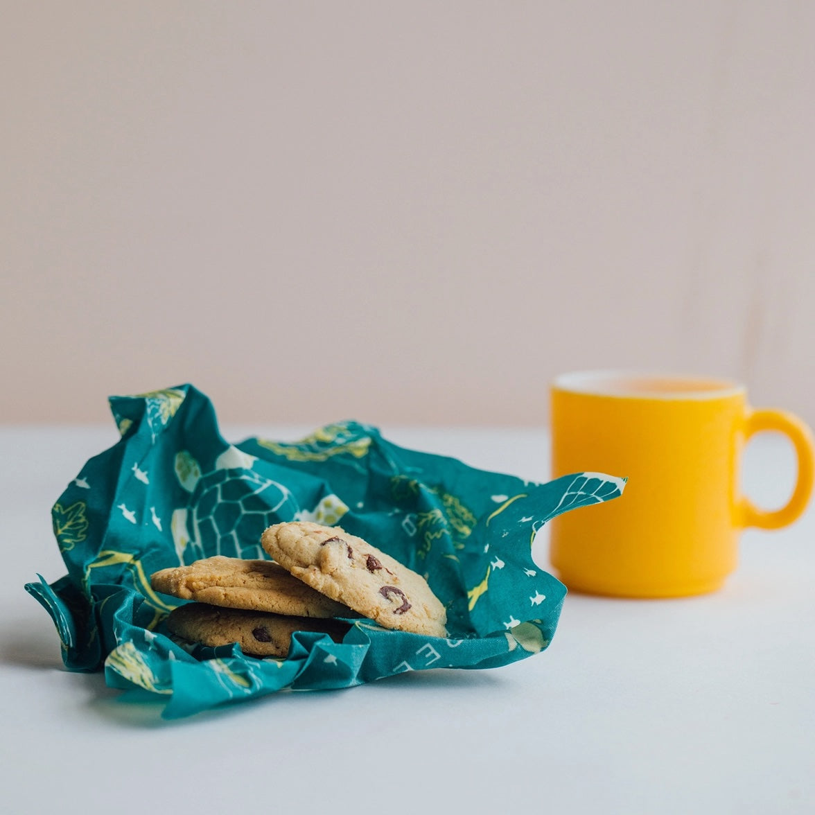 Three chocolate chip cookies wrapped in green Bee’s Wrap with a yellow mug in the background.
