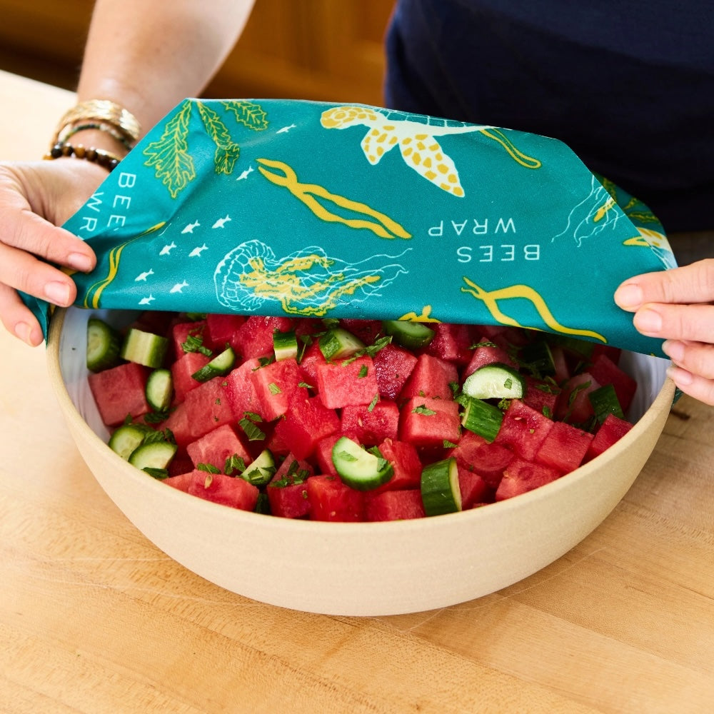 Hands holding a teal Bee’s Wrap with sea creatures, covering a bowl filled with chopped cucumbers and watermelon.