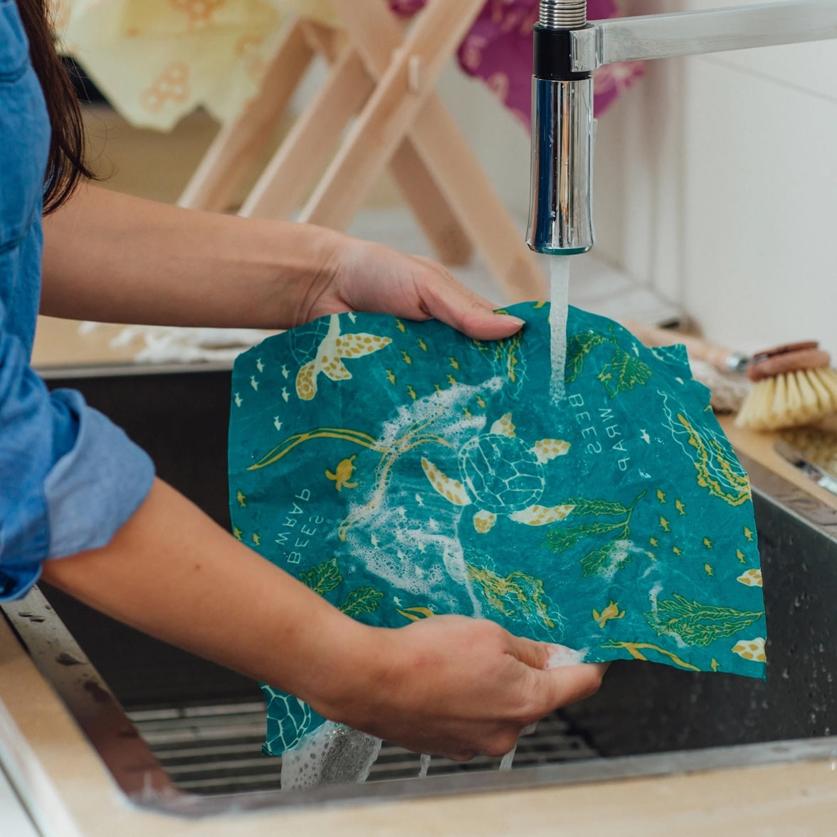 Hands washing a green Bee’s Wrap under running water at a kitchen sink.