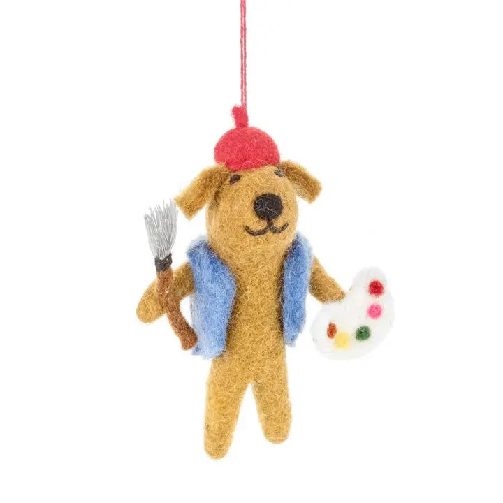 A handmade felt dog is shown dressed as an artist. It is wearing a red beret and a blue vest and is holding a paintbrush and a painter's palette.