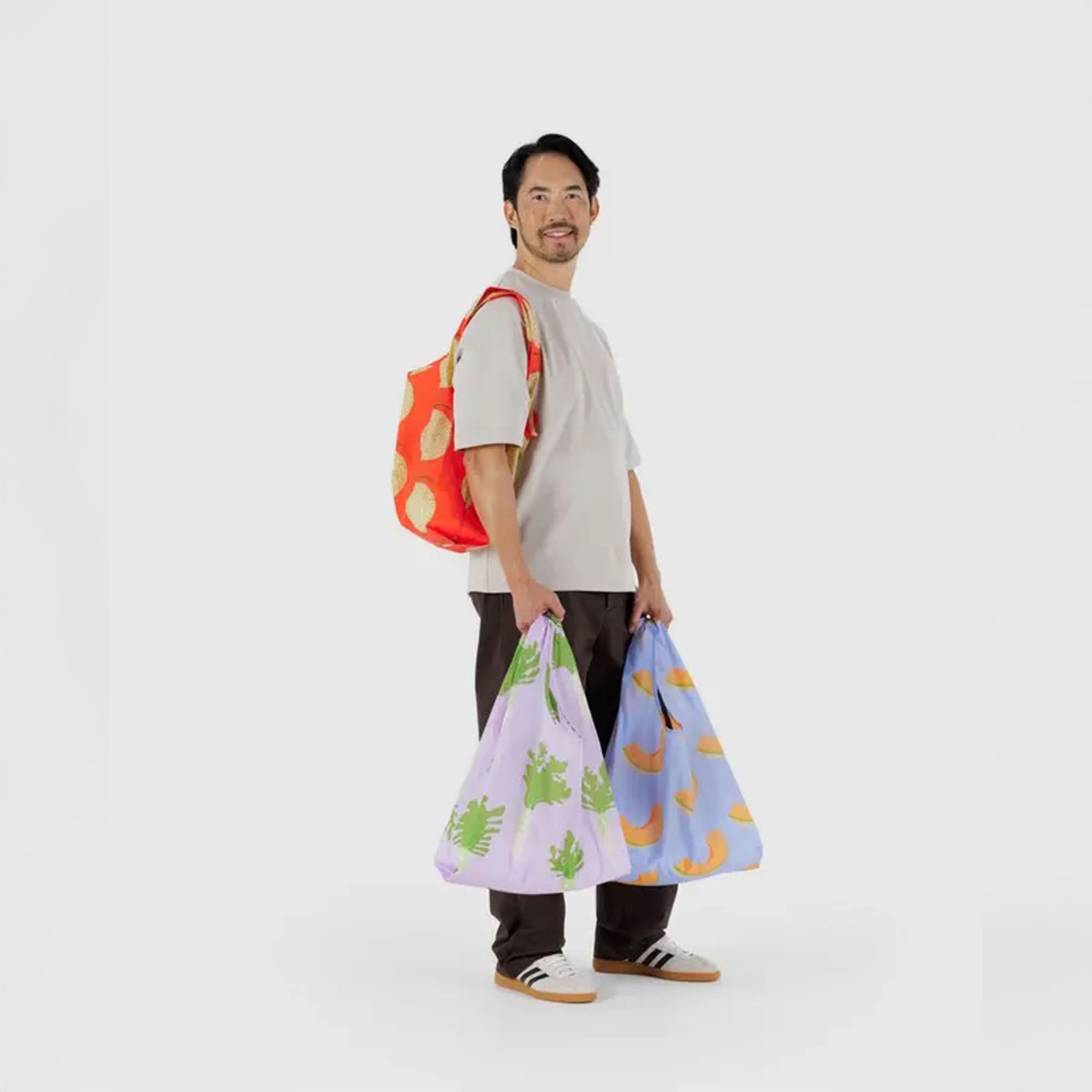 A person in a neutral-toned outfit carrying three BAGGU reusable bags in the “Produce Party” set, each bag showing off playful fruit patterns and everyday versatility.
