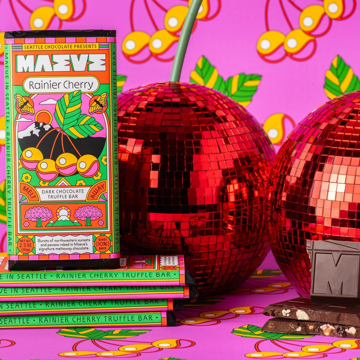 A Maeve “Rainier Cherry” truffle bar displayed on a pink festive surface next to cherry bright balls, highlighting the cherry-infused chocolate flavor.