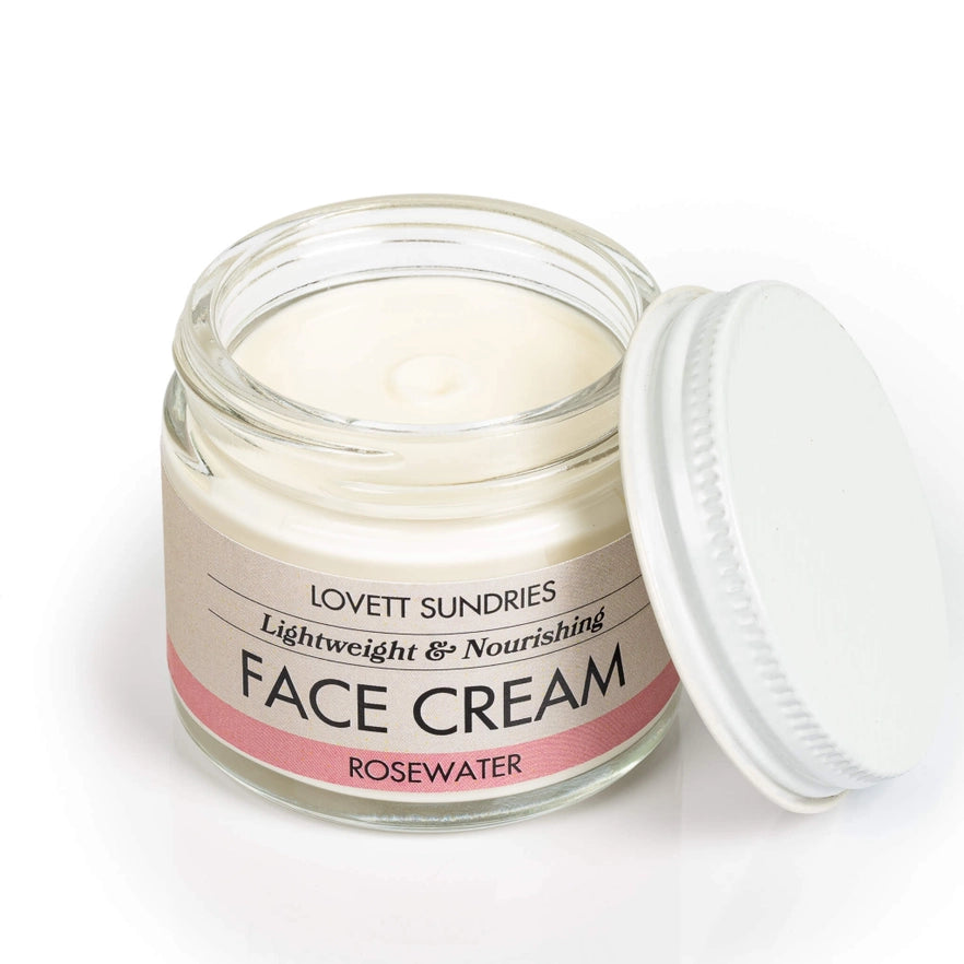 Open glass jar of rosewater face cream on a white surface, showing white cream inside and labeled metal lid beside it."