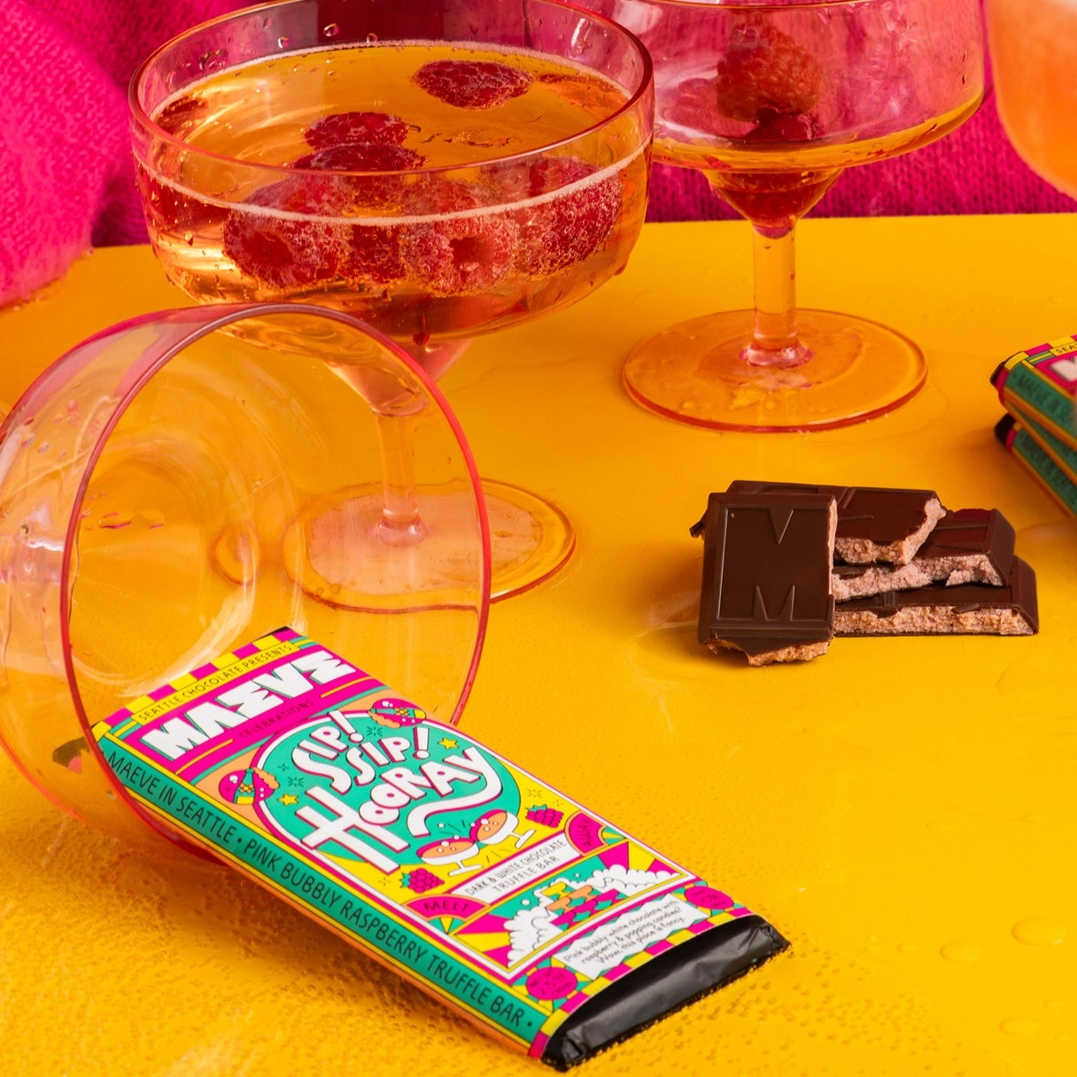 A close-up of a Maeve “Sip Sip Hooray” chocolate bar lying flat on a yellow surface, with party cups in the background.