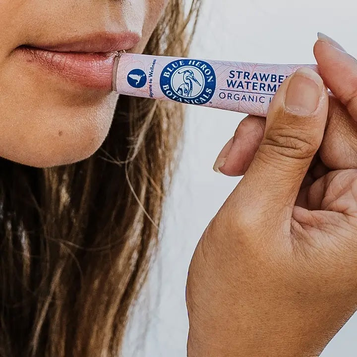 Woman applying Strawberry Watermelon Lip Therapy Balm, captured in soft natural light. Made with organic, nourishing ingredients and packaged in compostable, eco-friendly paperboard.