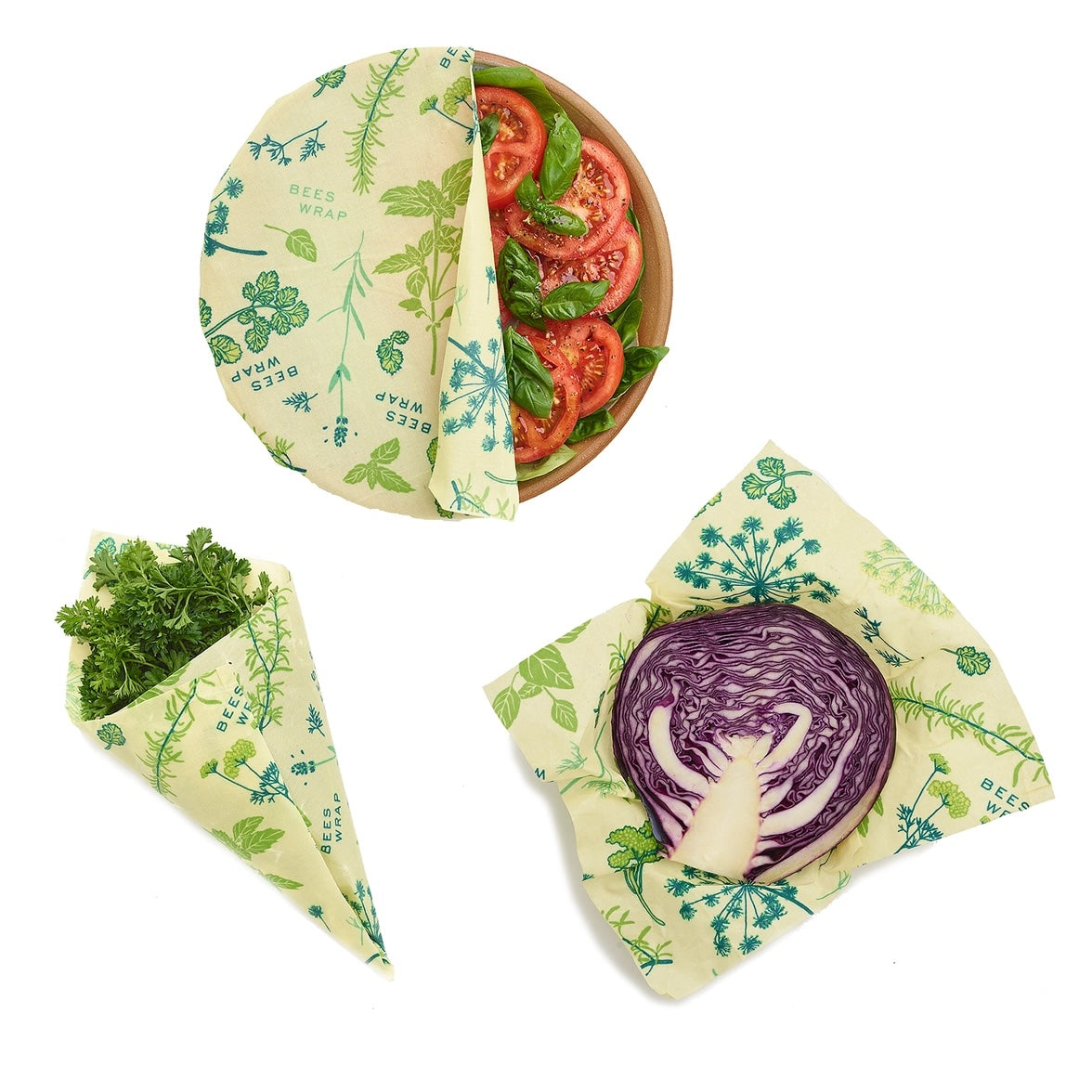Three beeswax wraps with green botanical prints covering fresh food: a bowl of sliced tomatoes and basil, a bundle of fresh herbs, and a halved purple cabbage.
