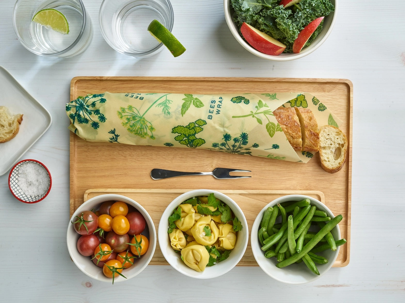 Wrapped sandwich with a botanical print Bees Wrap on a wooden tray, surrounded by bowls of kale salad, cherry tomatoes, and lime wedges.