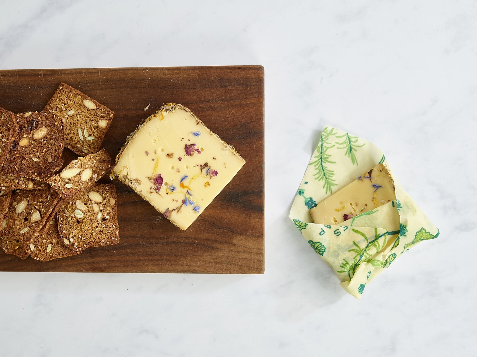A block of cheese with herbs and flowers wrapped in a yellow beeswax wrap with green botanical prints, placed on a white marble surface.
