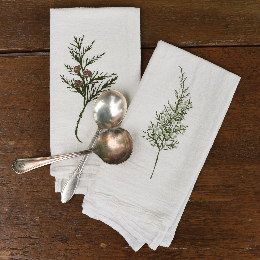 A lovely pair of white napkins is shown with two tarnished silver spoons resting on them. One towel has a charming green sprig with small pinecones, while the other features a delicate green leafy branch with tiny white buds.