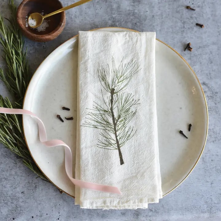 A charming white napkin with a green pine sprig design is folded on a white plate with a gold rim.