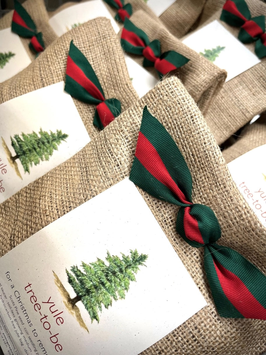 A close-up view of a collection of "yule tree to be" kits shows a repeating pattern of burlap sacks tied with red and green striped ribbons.