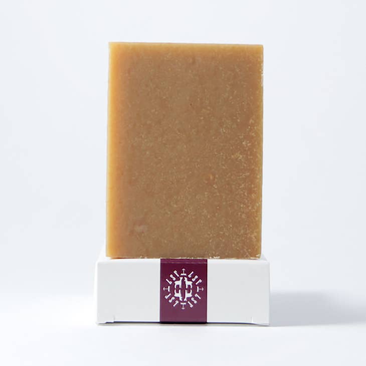 A light golden brown bar of soap is standing upright on a white box. The box has a dark red band around the center with a white logo on it.