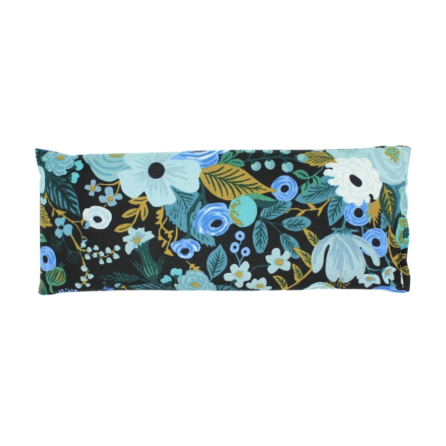 A rectangular lavender eye pillow with a dark background is covered in a floral pattern of blue white and green flowers and leaves.
