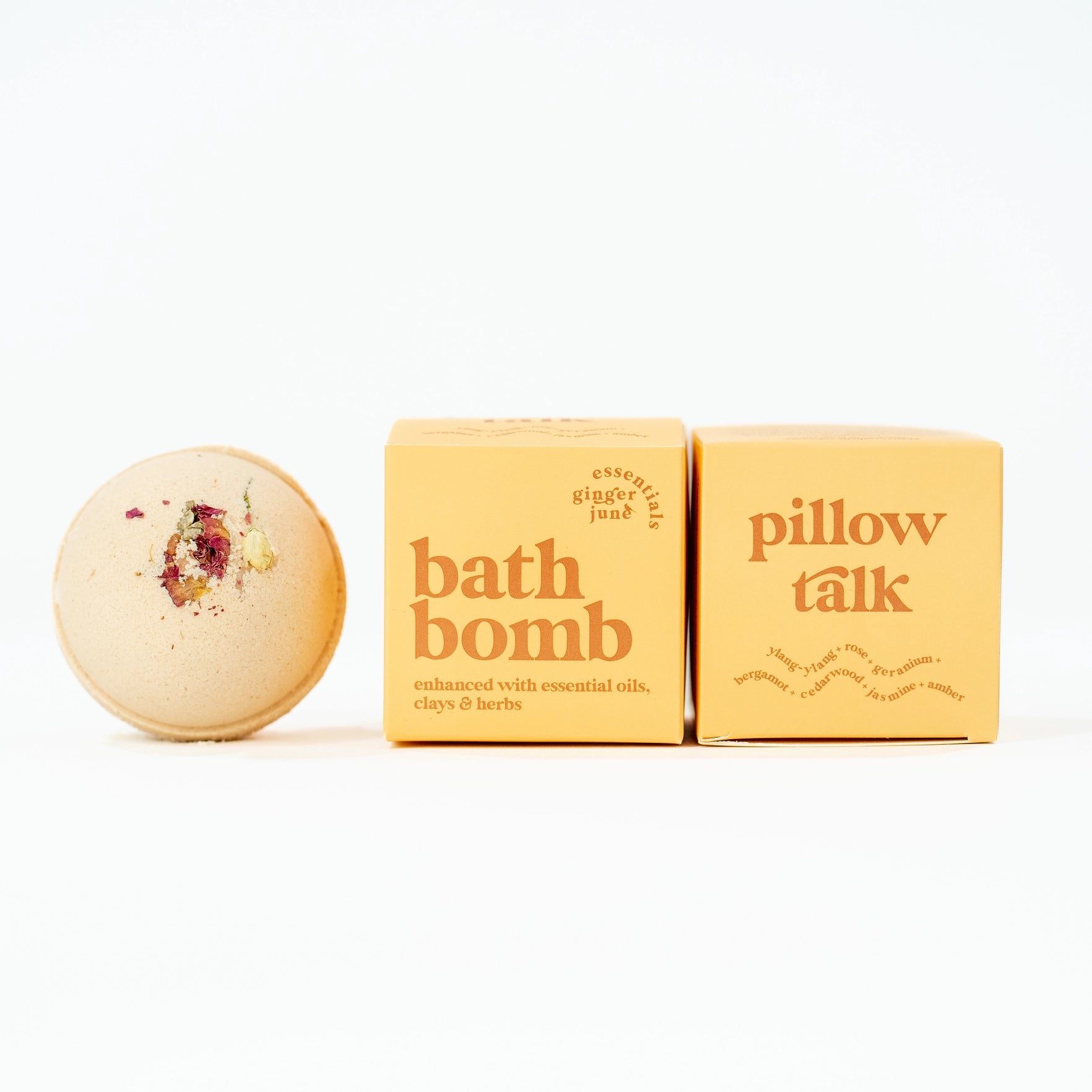 A light-colored bath bomb is shown next to two boxes. The bath bomb is topped with dried petals and herbs. The boxes are labeled ginger june essentials and pillow talk, with the essential oils ylang-ylang, rose, geranium, bergamot, cedarwood, jasmine and amber listed.