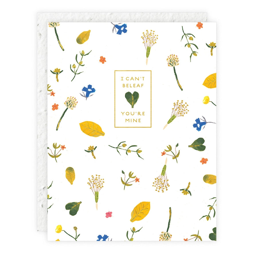 A seedling card featuring scattered green and yellow leaves and wildflowers on a white background, with gold text: "I CAN'T BELEAF YOU'RE MINE."