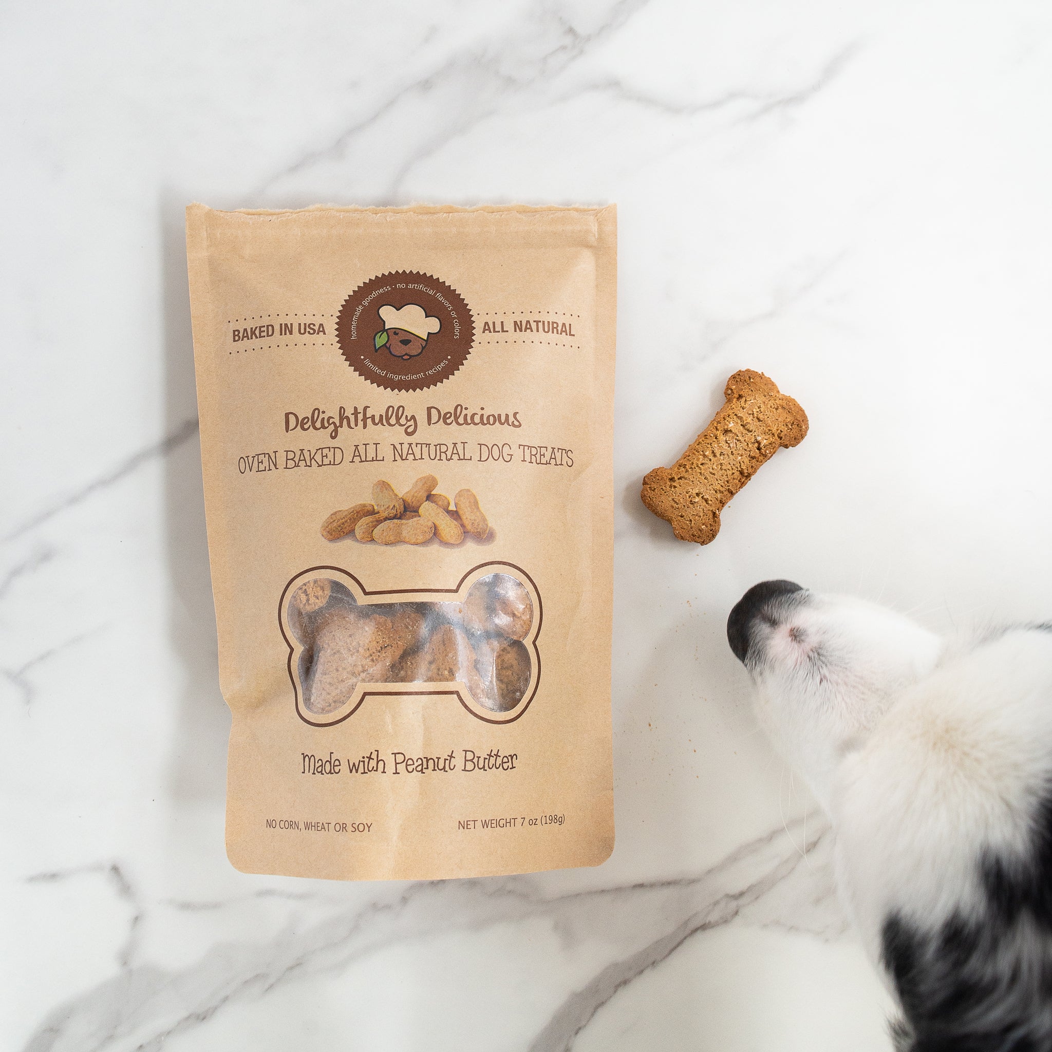 A bag of Delightfully Delicious dog treats is shown on a white marble surface. The bag is labeled "Made with Peanut Butter". A bone-shaped biscuit sits next to the bag, while a black and white dog's snout is visible in the corner, looking at the treat.