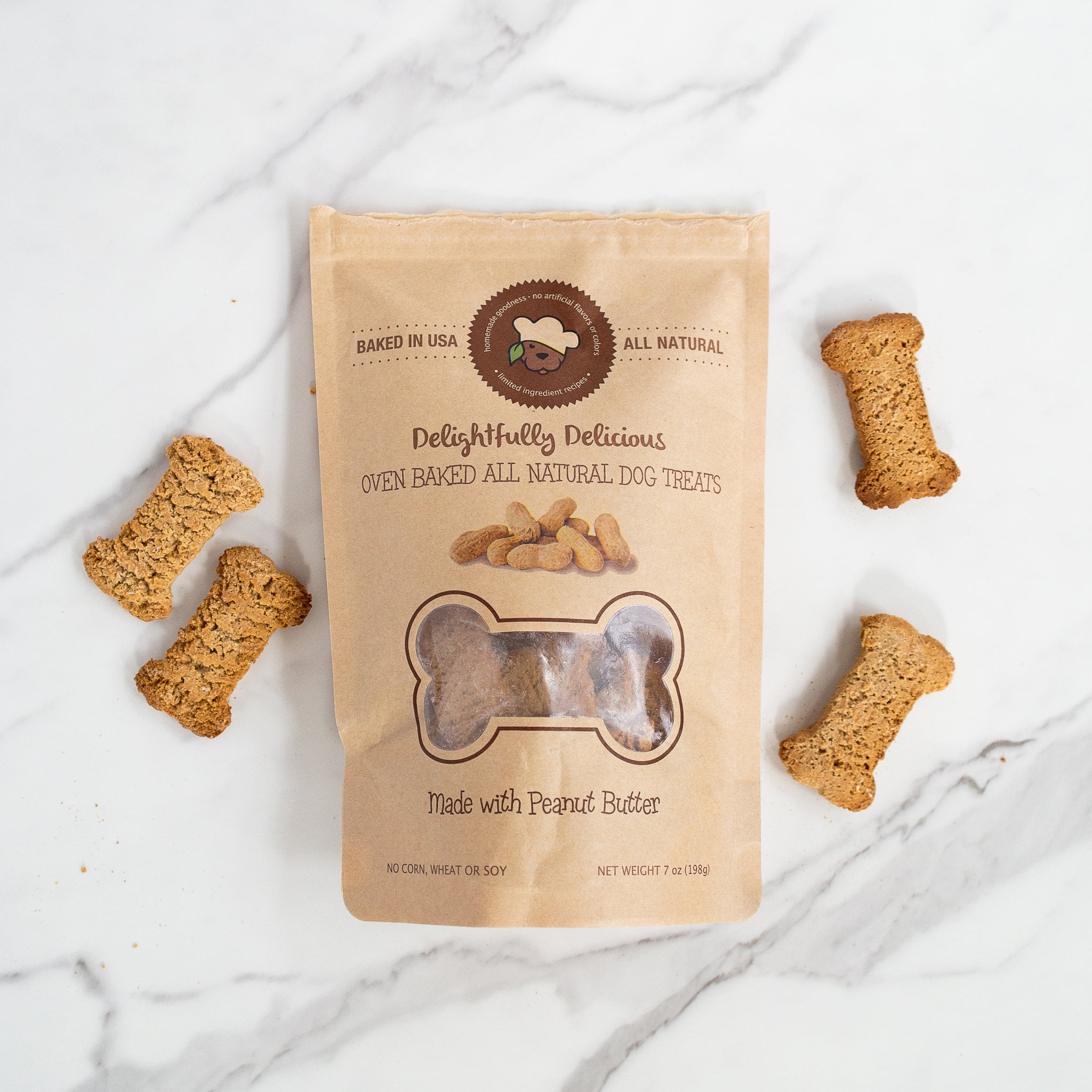 A bag of dog treats is shown on a white marble surface with four bone-shaped biscuits placed around it. The bag, labeled Delightfully Delicious and "Made with Peanut Butter", it has a bone-shaped cutout revealing the treats inside.
