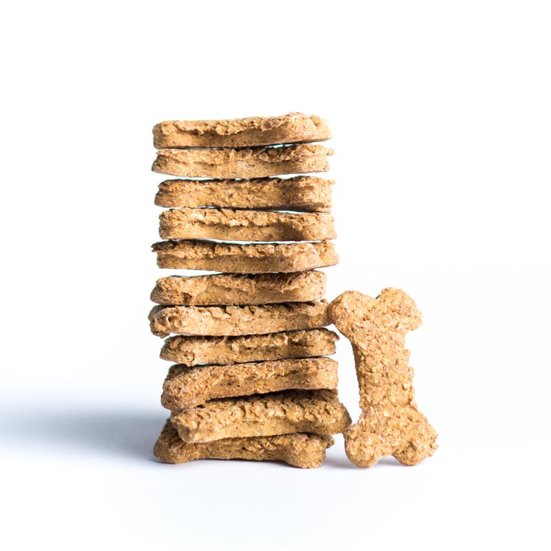 A stack of bone-shaped dog treats is shown against a white background, with a single biscuit standing on its end to the side. The treats are a light brown color with an oatmeal-like texture.