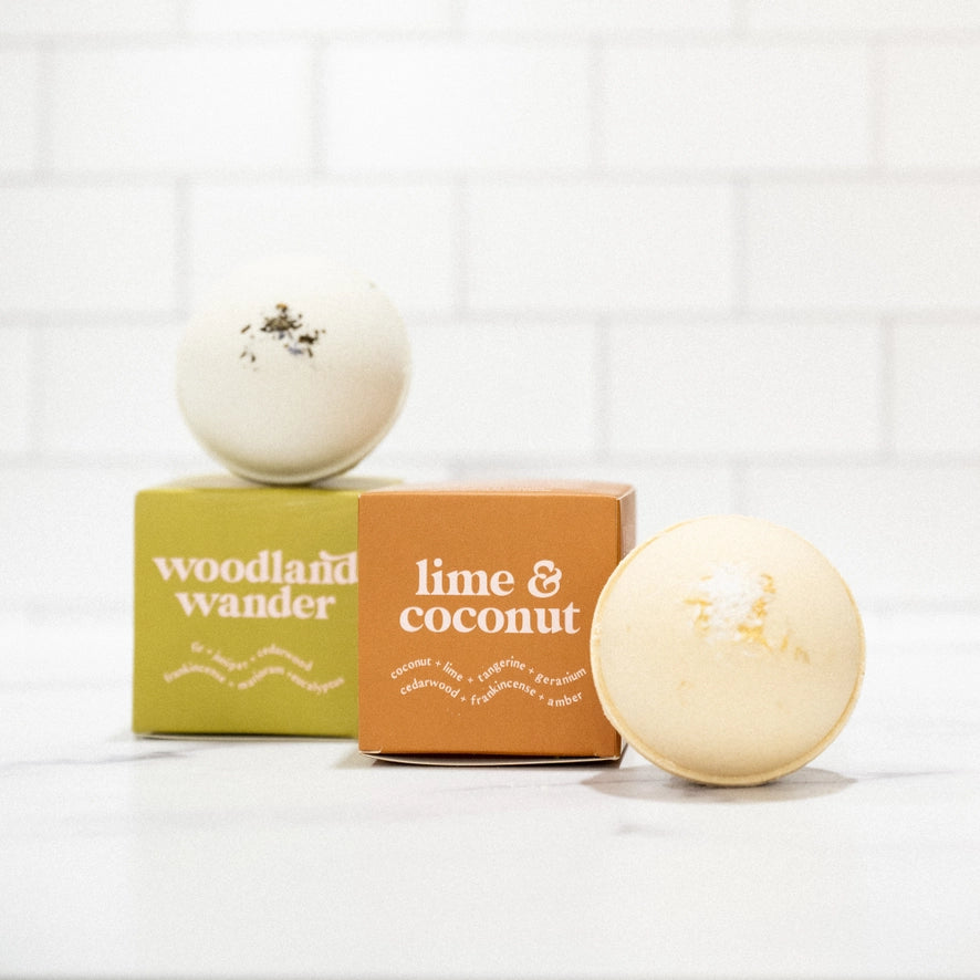 Two bath bombs and two boxes are shown against a white brick background. The bath bomb on the left sits on a box labeled woodland wander. A second bath bomb and an orange box labeled lime & coconut are on the right.