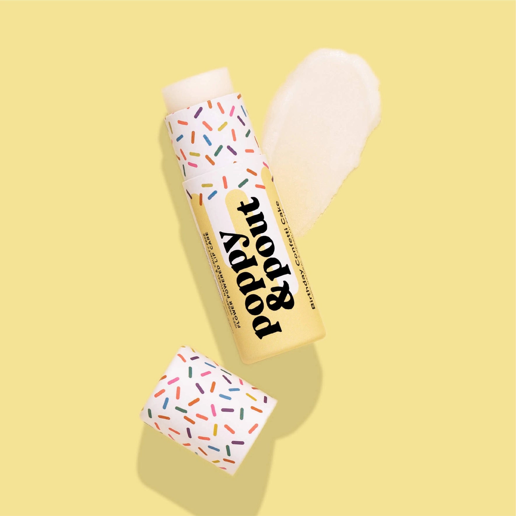 An open tube of poppy & pout lip care is shown on a light yellow background. The tube and its cap are decorated with a colorful confetti pattern. The lip care is labeled "Birthday Confetti Cake," and a swatch of the creamy white product is next to the tube.