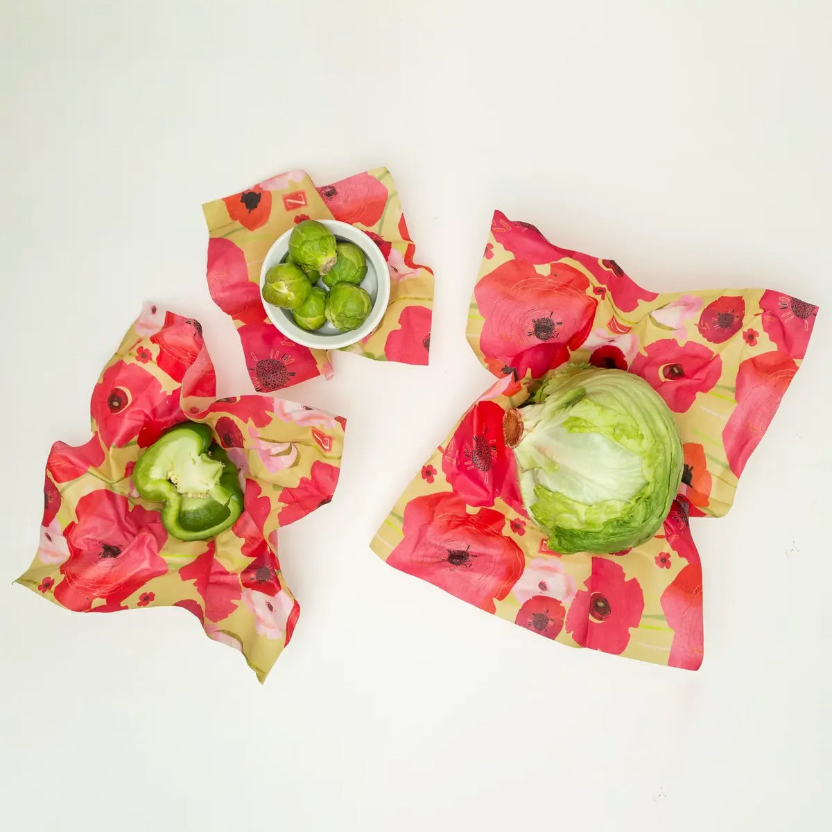 Three reusable beeswax food wraps in colorful flower patterns holding fresh vegetables.