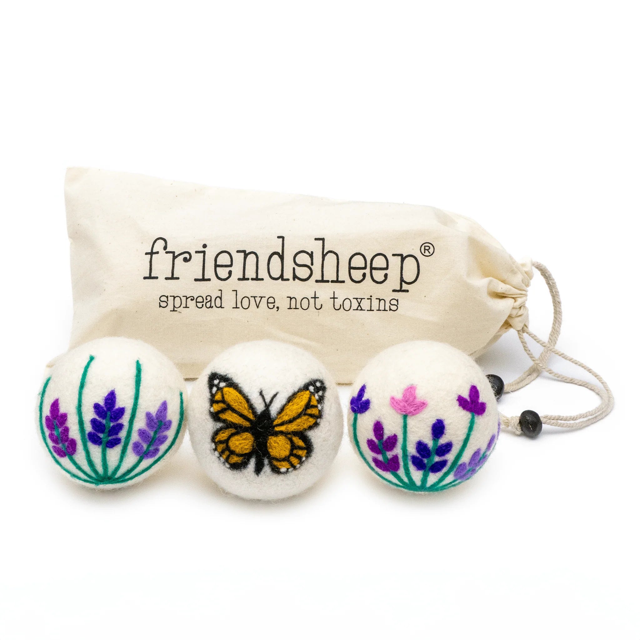 A set of three Friendsheep sustainable goods wool dryer balls are shown decorated with a cute butterfly and two floral designs. They are sitting in front of their white canvas drawstring bag.