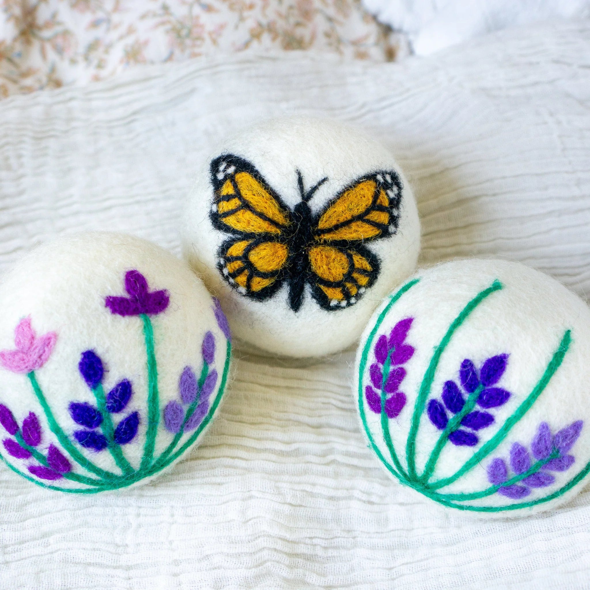 A set of three Friendsheep sustainable goods wool dryer balls, decorated with a cute butterfly and two different purple floral designs are resting on a rumpled white cloth.