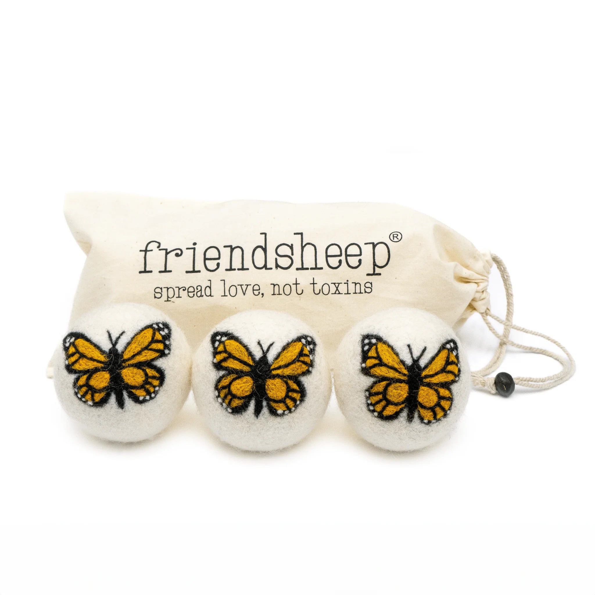 A set of three Friendsheep sustainable goods wool dryer balls are shown, each decorated with a cute butterfly. They are sitting in front of their white canvas drawstring bag.
