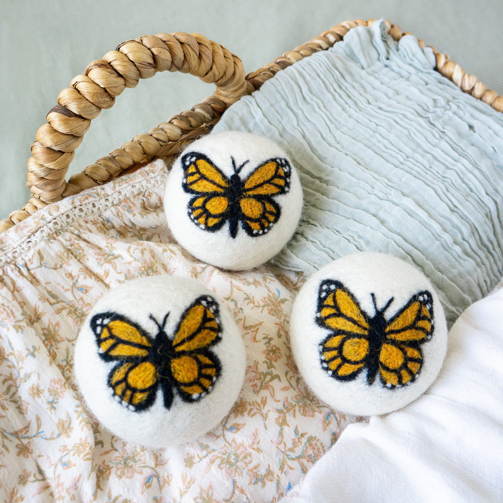 A set of three adorable Friendsheep sustainable goods wool dryer balls, each decorated with a cute monarch butterfly, are nestled in a wicker basket on a floral-patterned cloth.