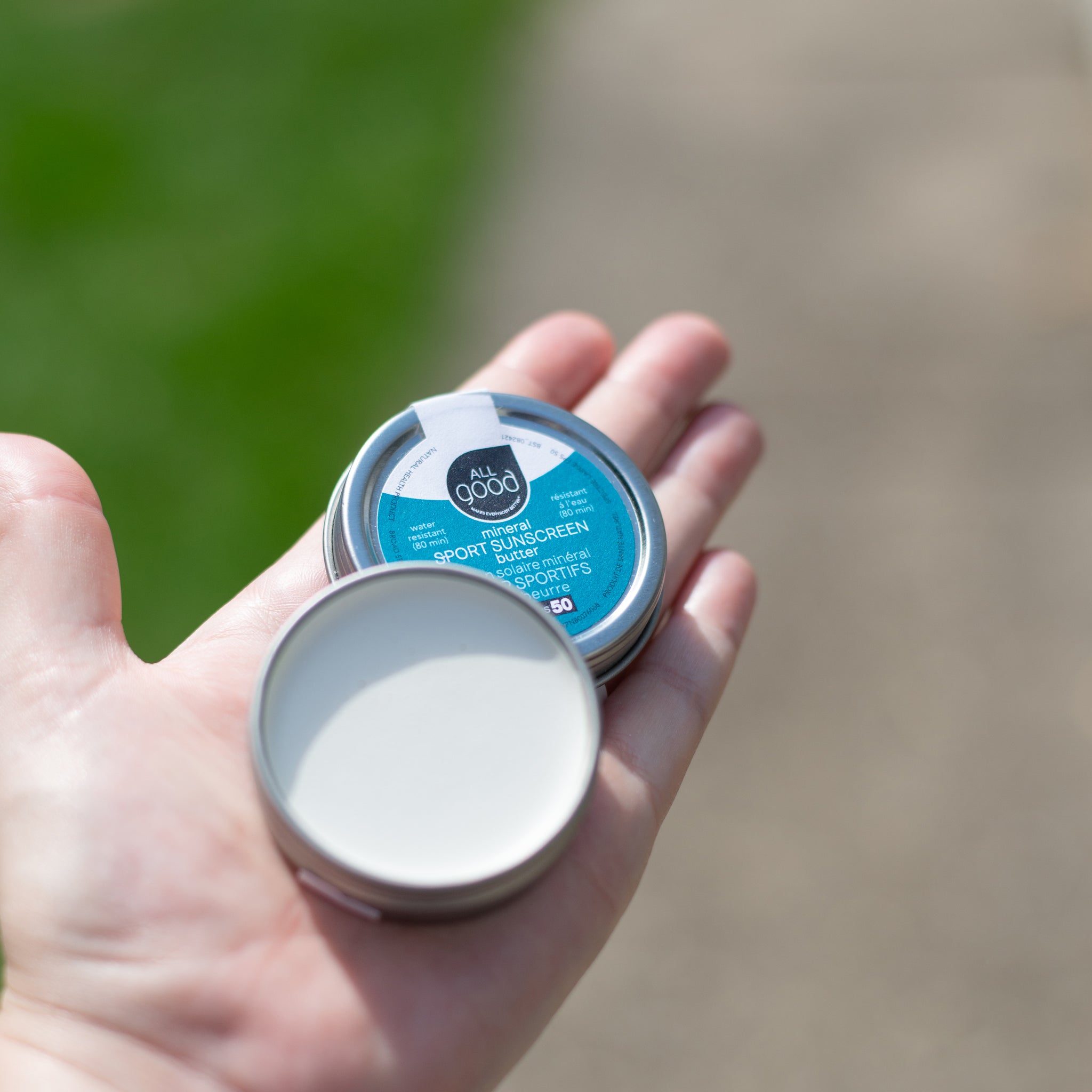 A hand holds an open tin of All Good mineral SPORT SUNSCREEN butter over a blurry green background. The lid is off and the solid sunscreen is visible.