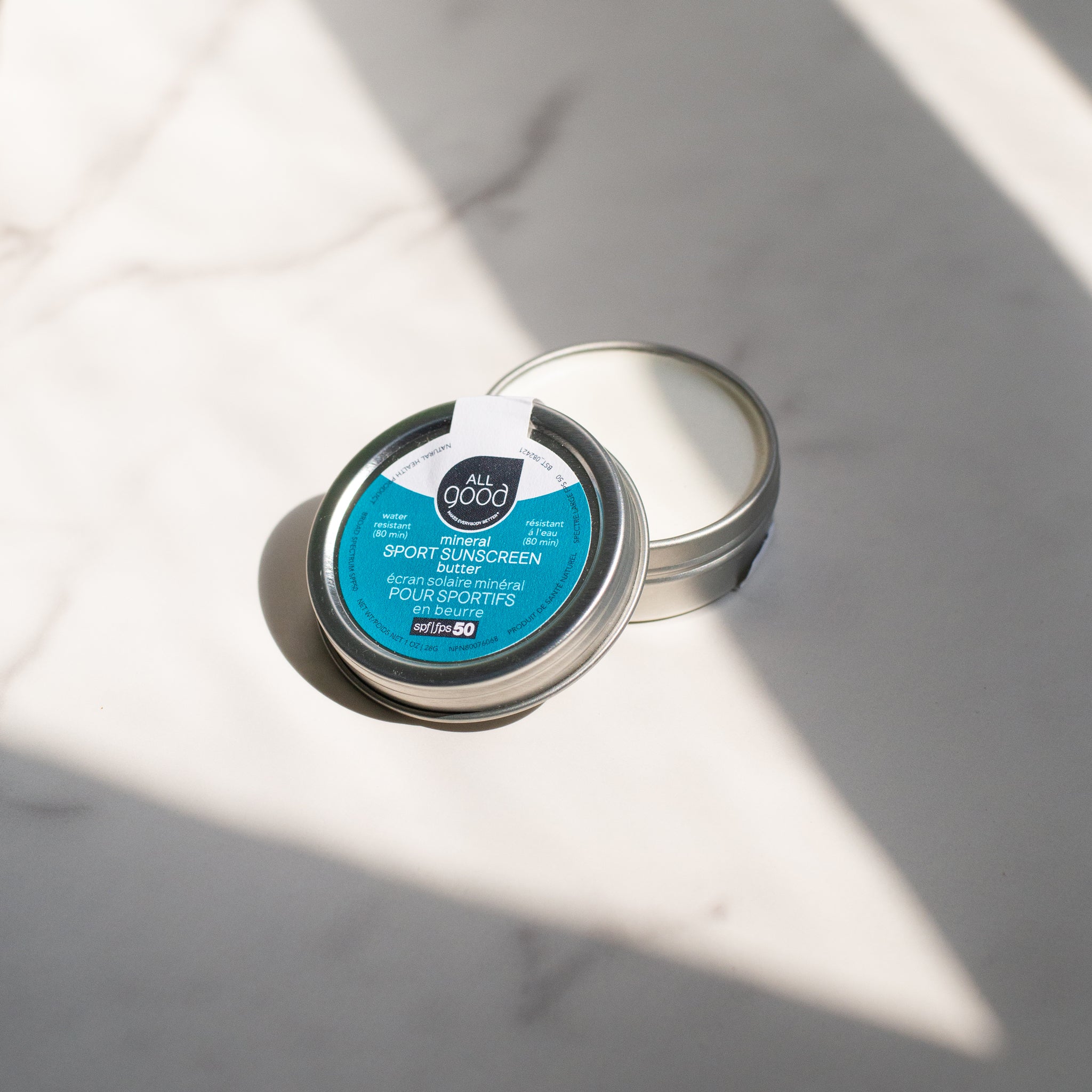 A small open tin of All Good Mineral Sport Sunscreen Butter SPF 50 rests on a white marble surface. The tin is silver with a blue label and the white cream inside is visible.