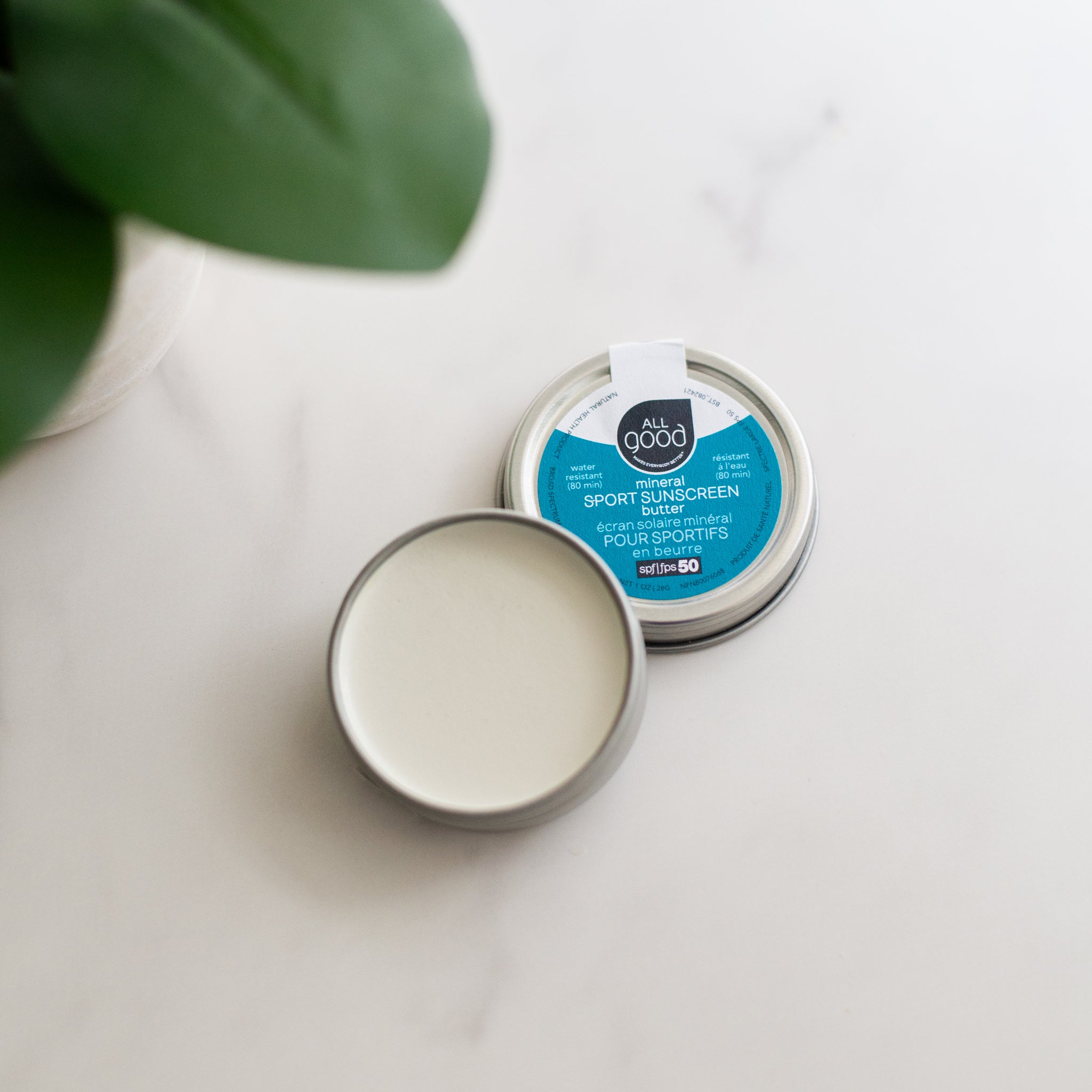 An open tin of All Good mineral SPORT SUNSCREEN rests on a white marble surface, next to its lid. A green plant is in the background.