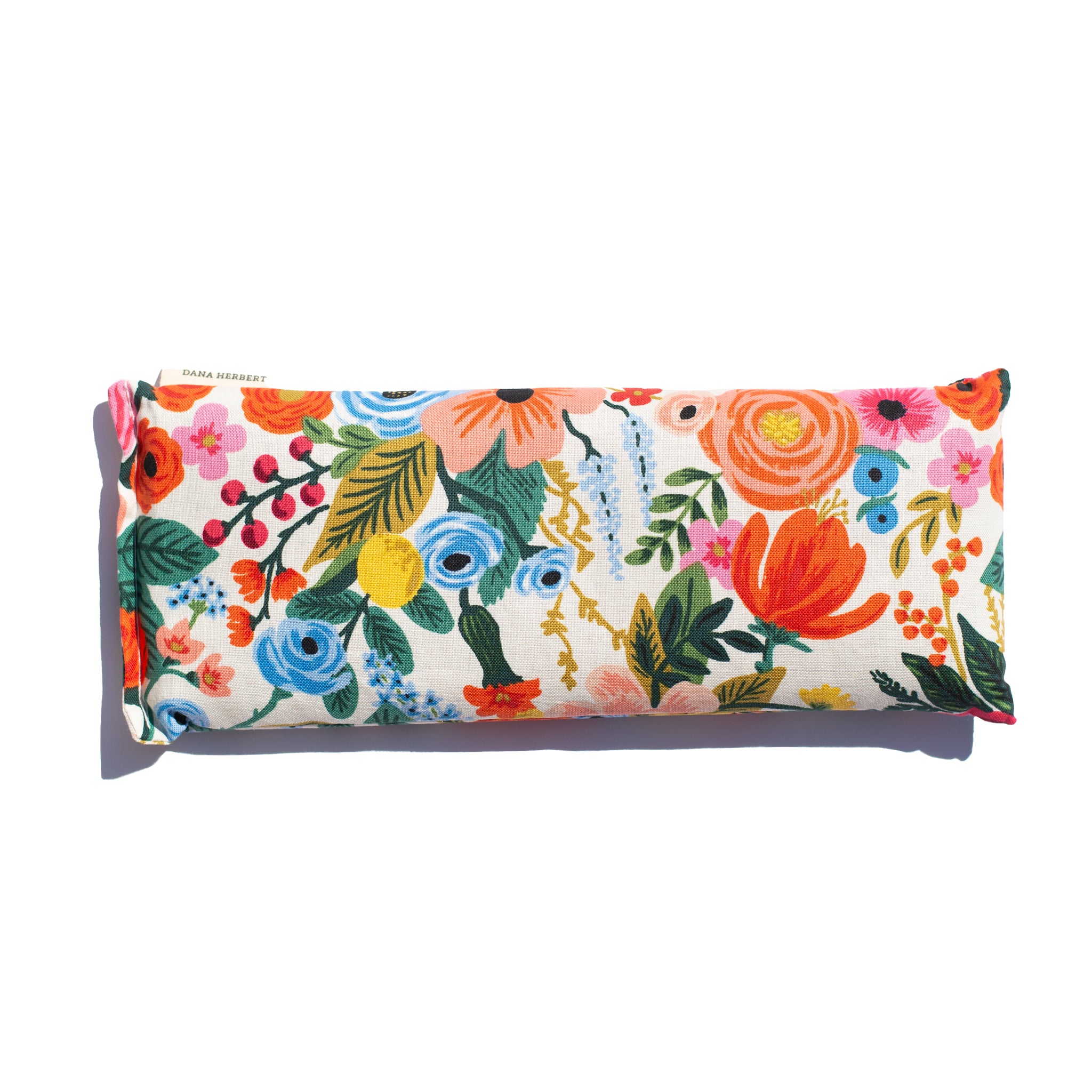 A rectangular lavender relaxation eye pillow has a vibrant floral pattern with orange pink and blue flowers. The brand "Dana Herbert" is on a small tag.