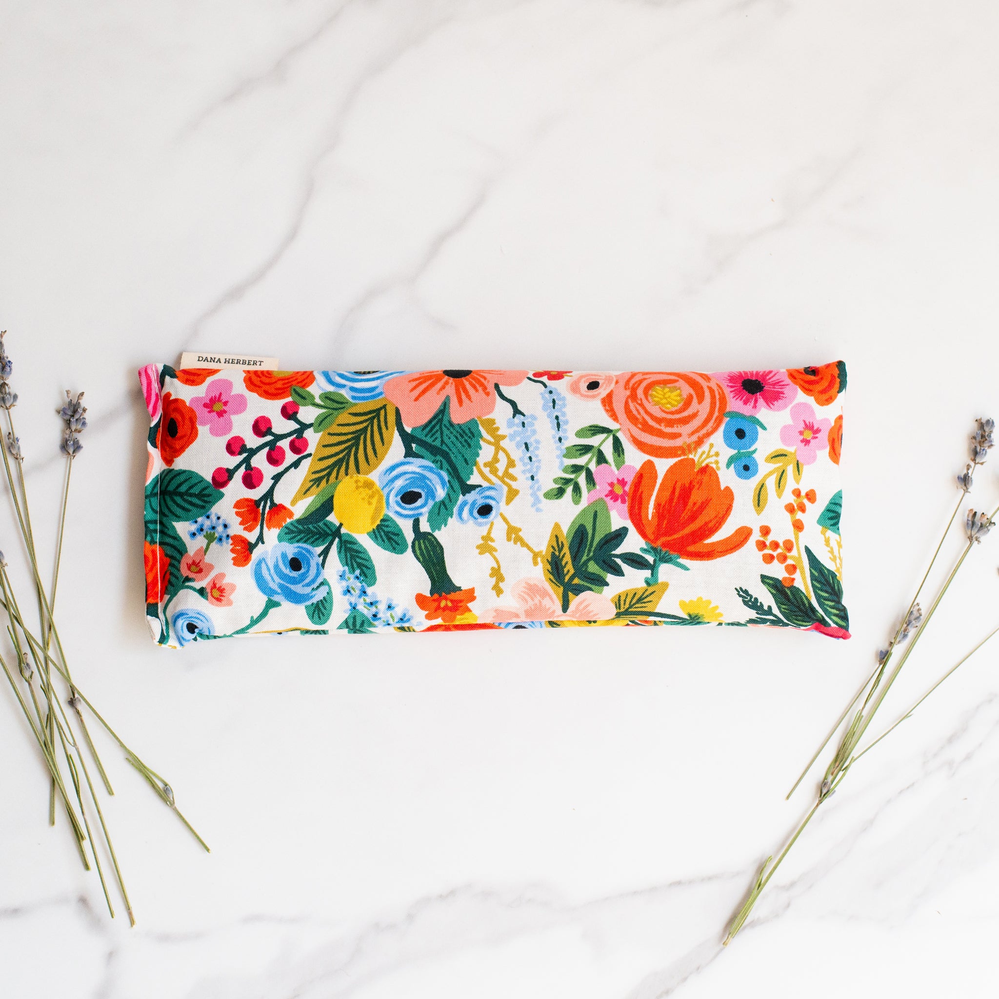 A floral patterned lavender eye pillow lies on a white marble surface with dried stalks of lavender on either side.