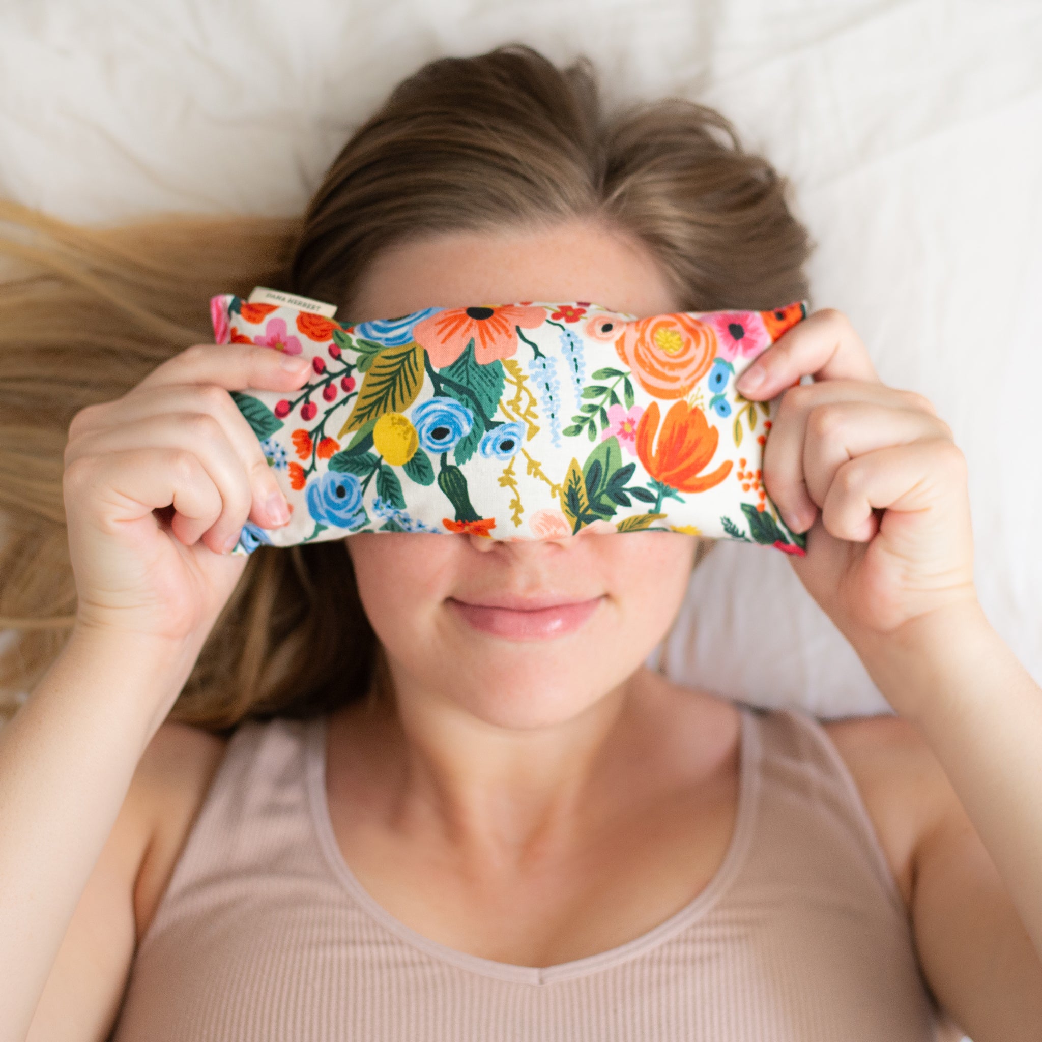 A woman lies on a bed holding a rectangular floral-patterned lavender relaxation eye pillow over her eyes. She is smiling faintly and her blonde hair is fanned out on the pillow.