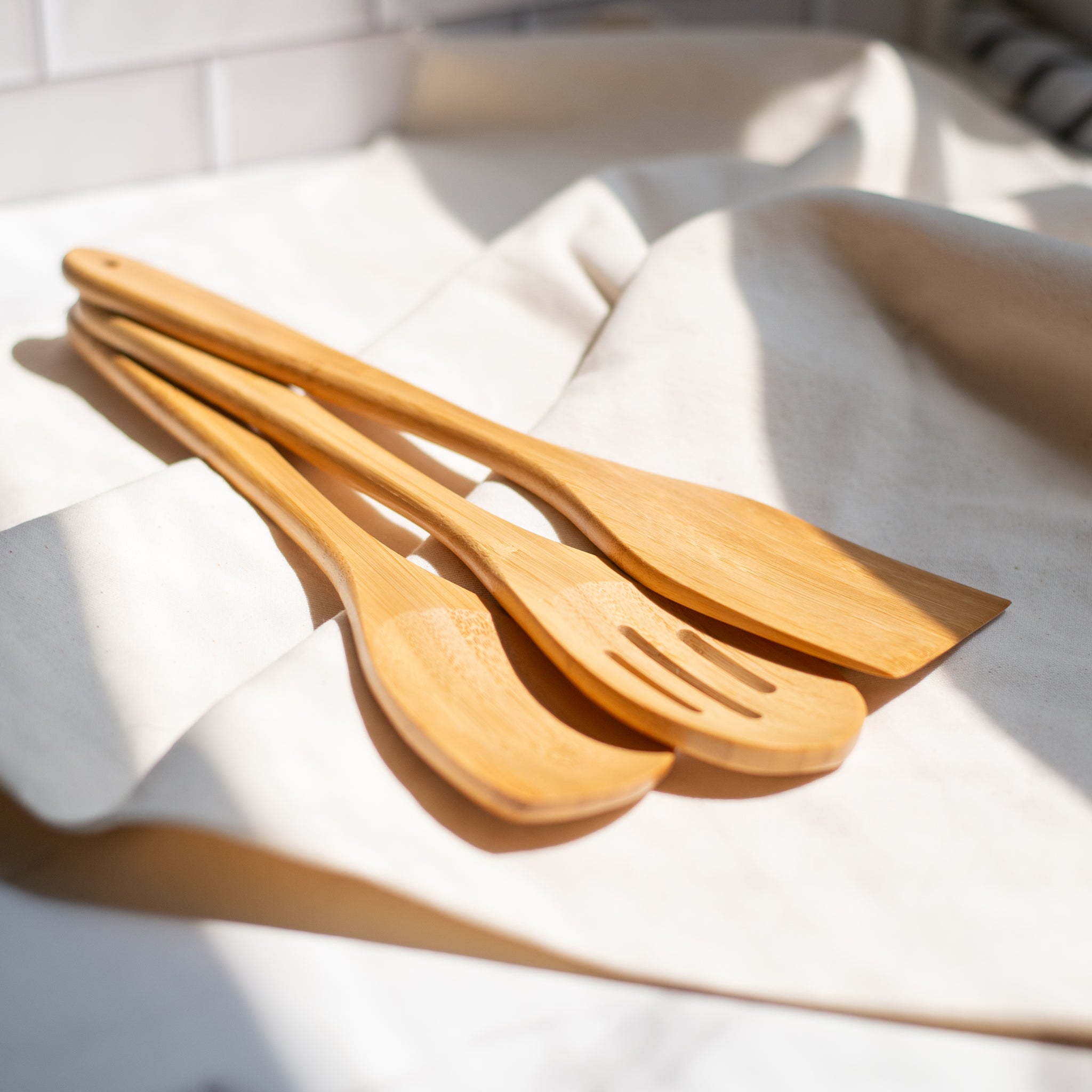 Three bamboo cooking utensils resting on a beige linen cloth, showing natural wood grain and smooth finish. Sustainable, organic bamboo kitchen tools with no glues or lacquer.