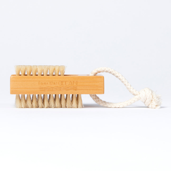 A bamboo nail brush with two sets of bristles. The handle has a Free the OCEAN logo etched into it and a braided rope loop is attached to one end.
