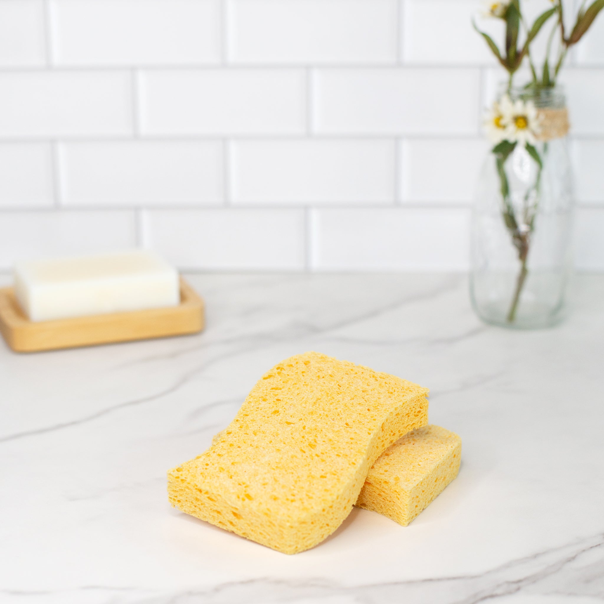 Two natural plant cellulose biodegradable kitchen sponges in a marble surface with some white flowers in a jar.