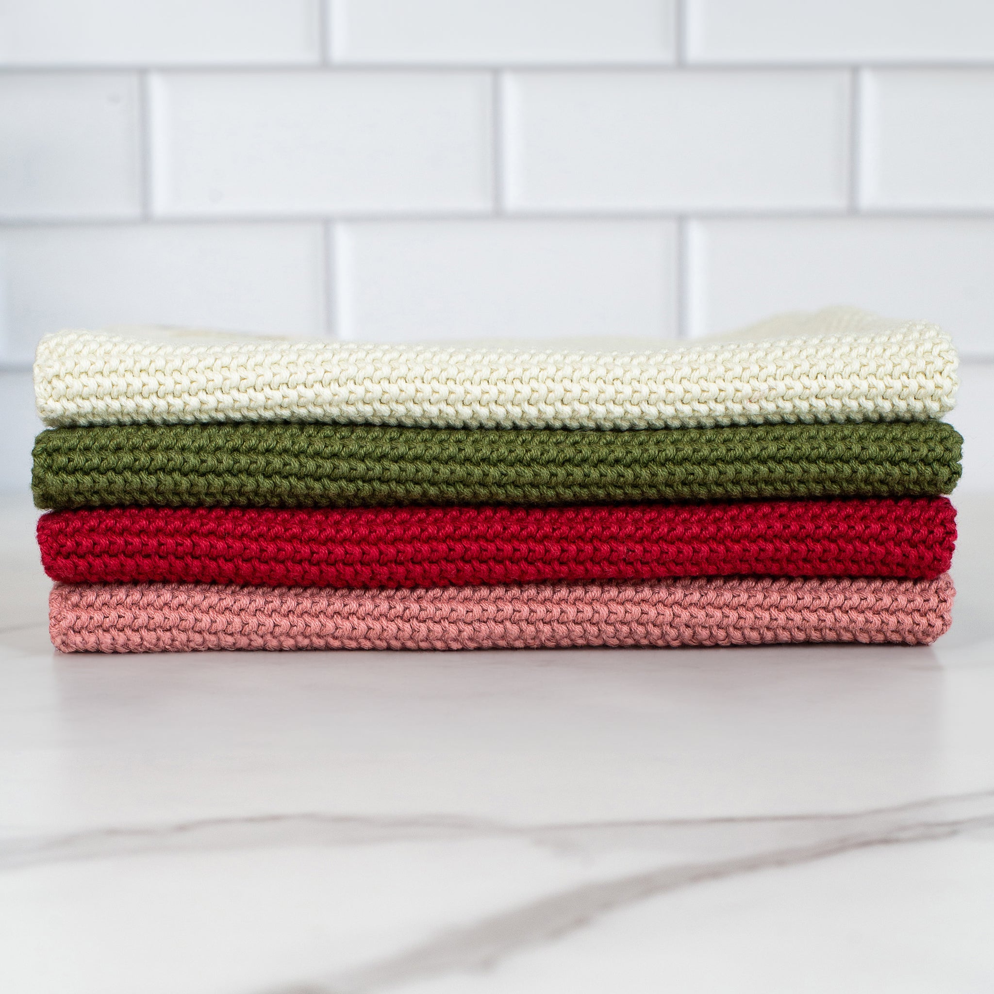 A stack of four rectangular cleaning cloths with a waffle texture. From top to bottom they are light cream, olive green, dark red and muted rose.