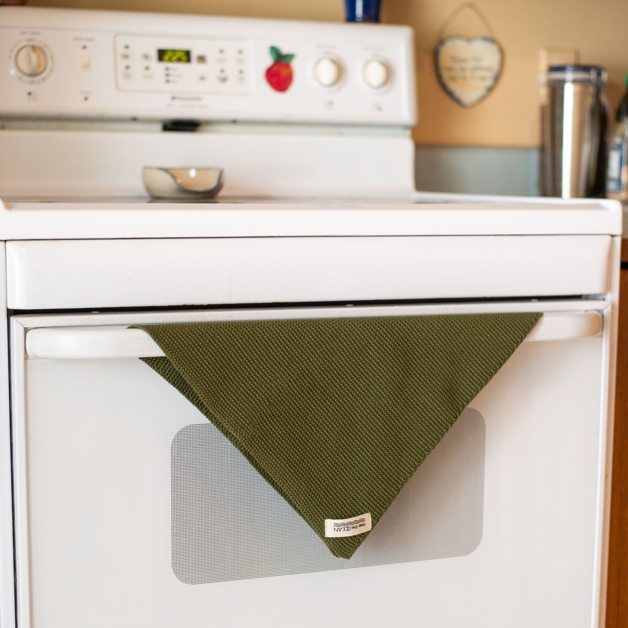 A dark olive green cleaning cloth with a waffle texture is folded into a triangle and draped over the handle of a white oven. A small white tag is visible at the bottom of the triangle.
