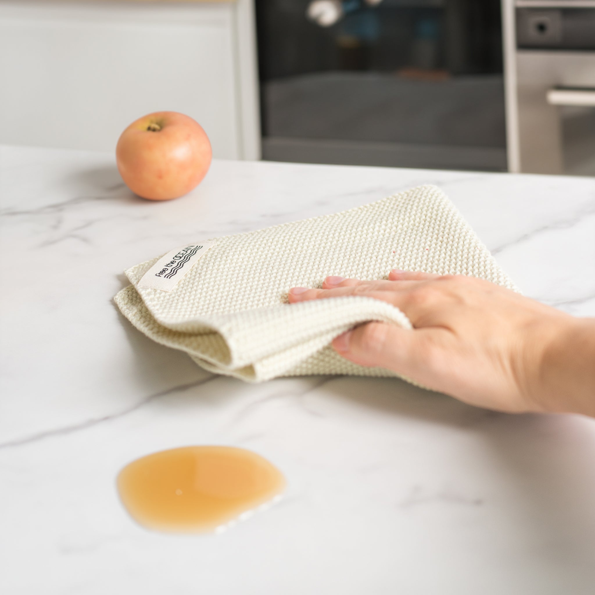 A light cream colore dishcloth with a waffle texture is being held by a hand about to wipe up a small puddle of an amber liquid on a white marble countertop.