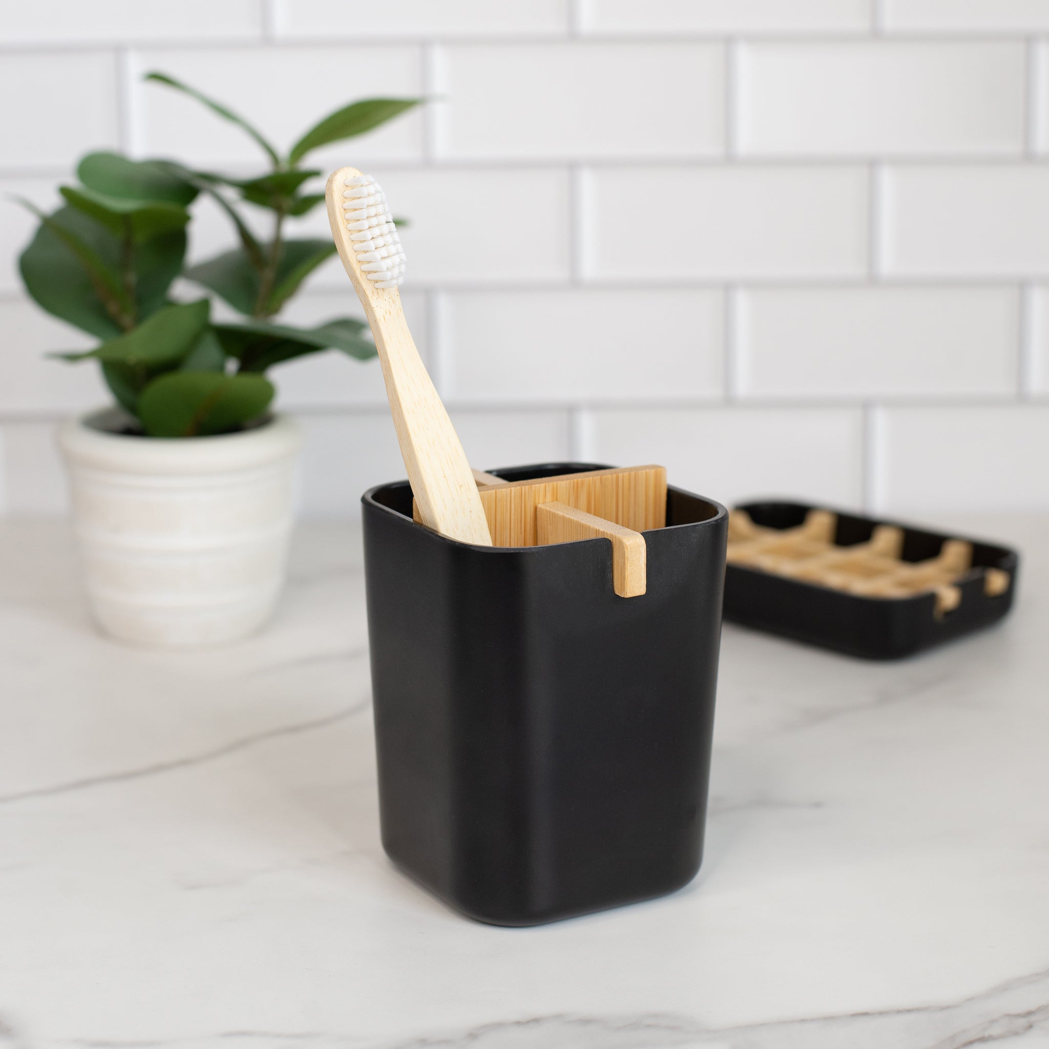 A matte black rectangular toothbrush holder with a removable bamboo divider. A bamboo toothbrush with white bristles is placed in one of the holder's sections. Another bamboo soap dish rests on its black base nearby.