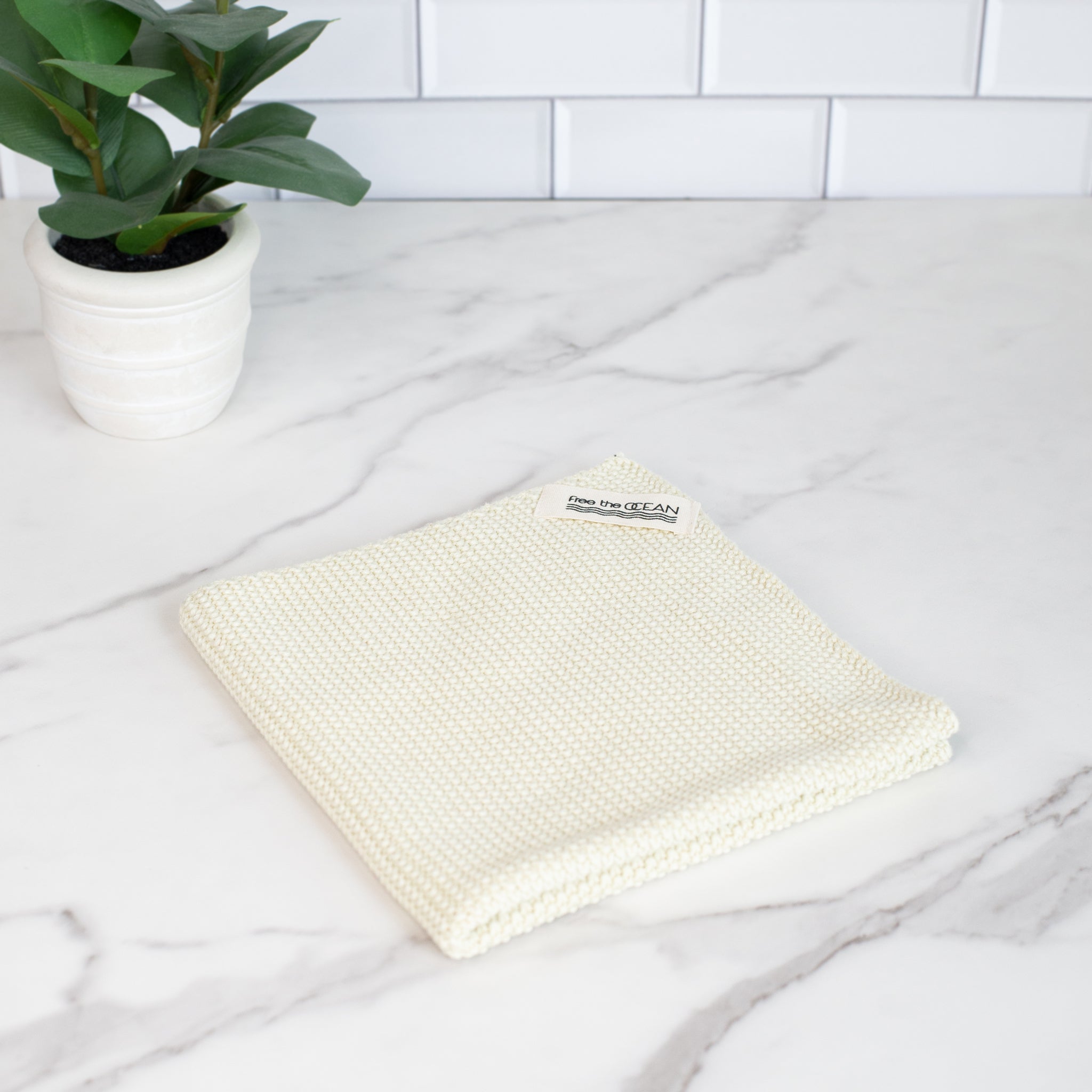 Light beige, textured cleaning cloth with a small tag that reads "Free the Ocean." The cloth is folded neatly and rests on a white marble surface.